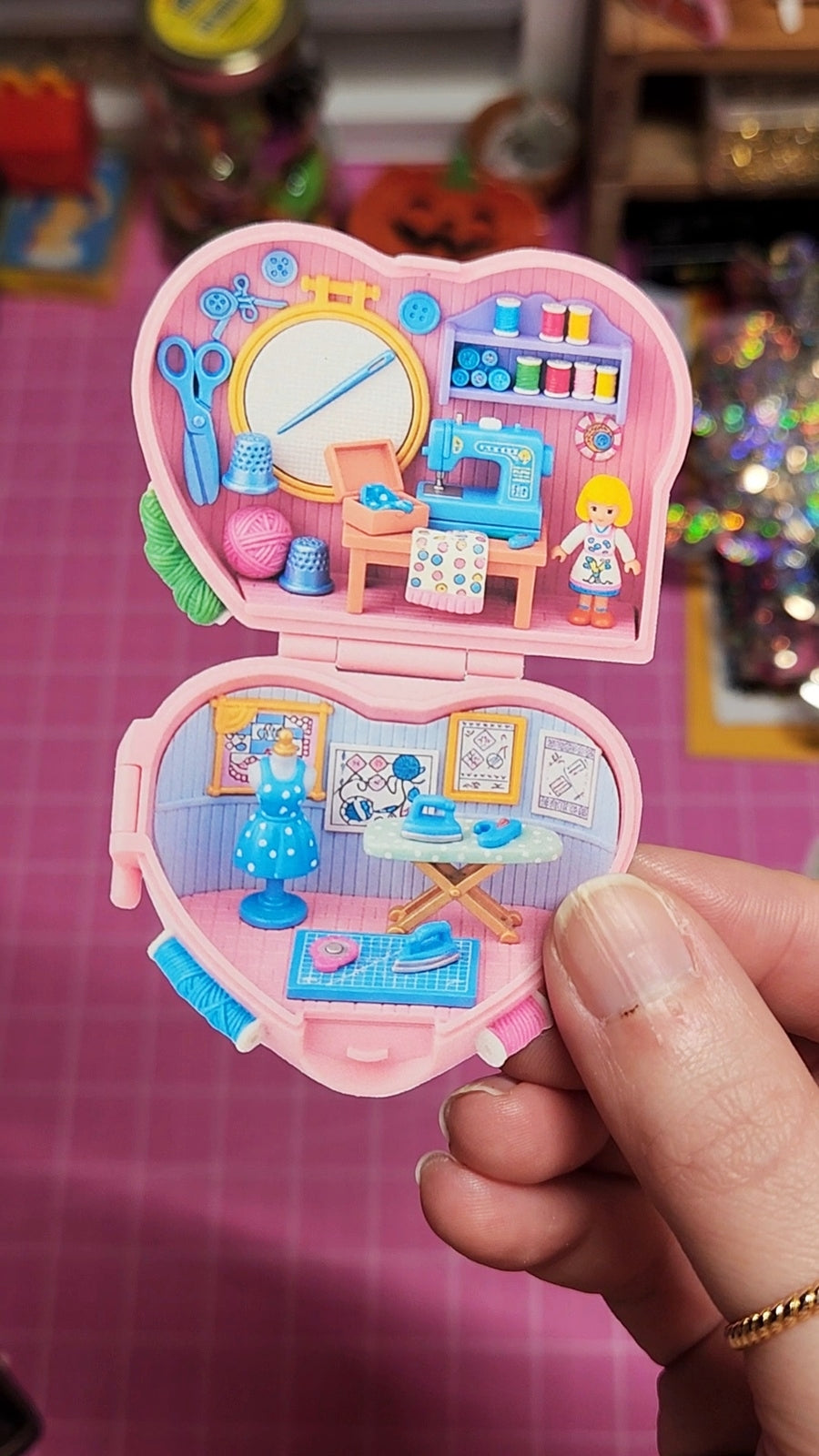 Heart-shaped toy with a miniature room scene held by a hand on a pink surface.