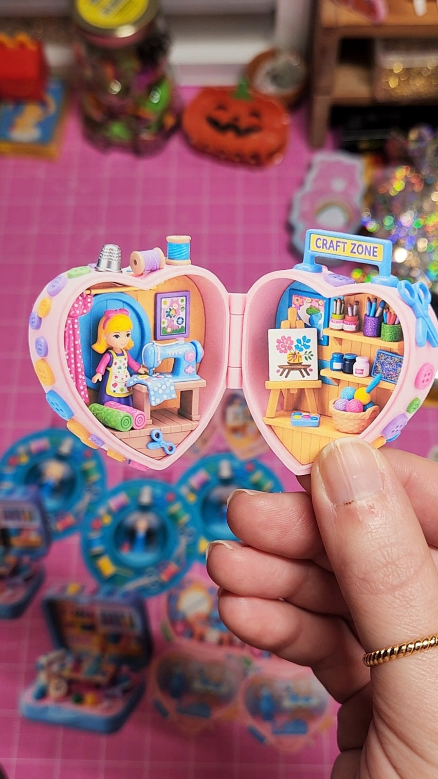 Heart-shaped toy with a doll and craft supplies on a pink grid background