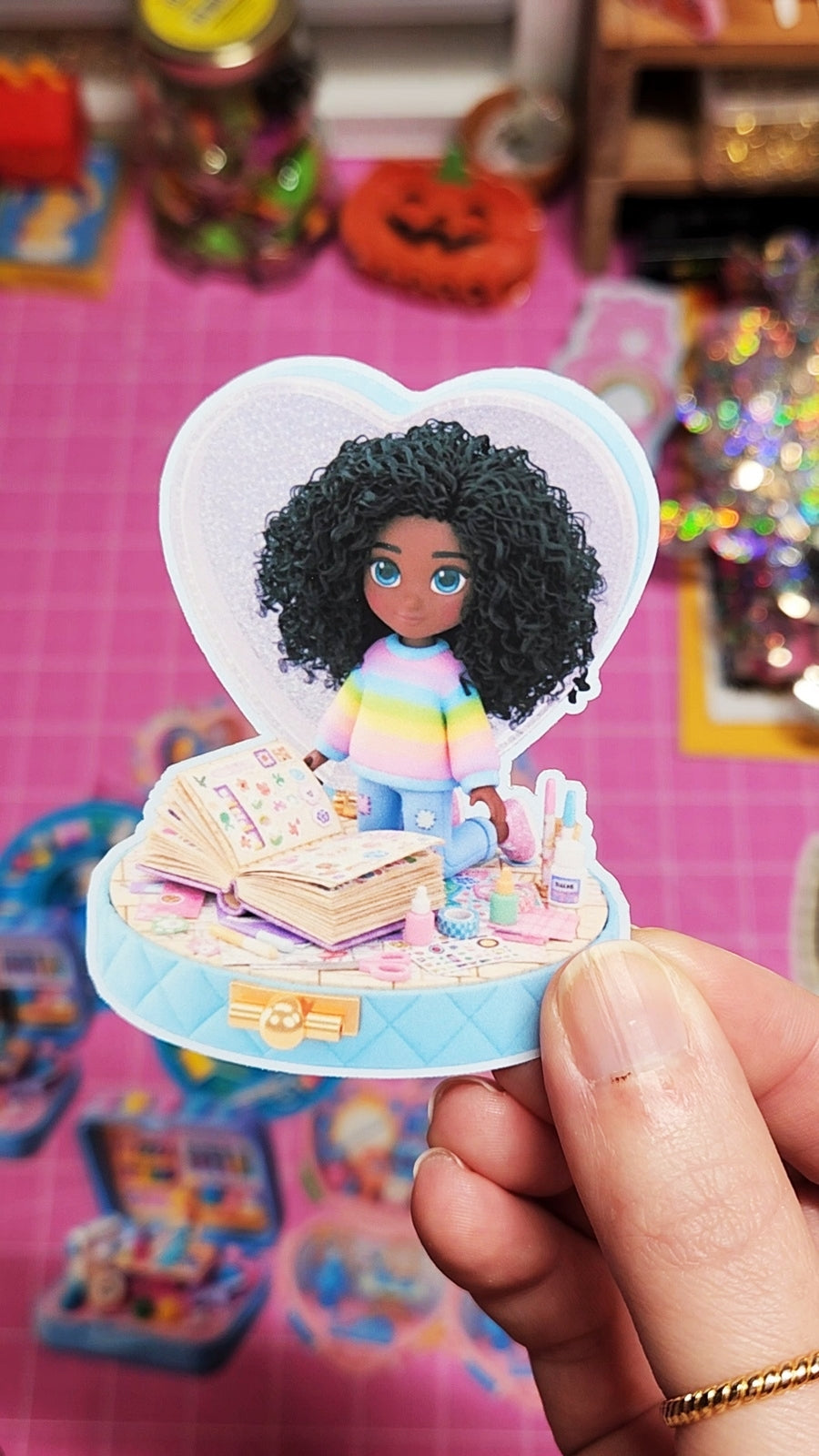 Small figurine of a person with curly hair in a heart-shaped display case, held against a colorful, blurred background.