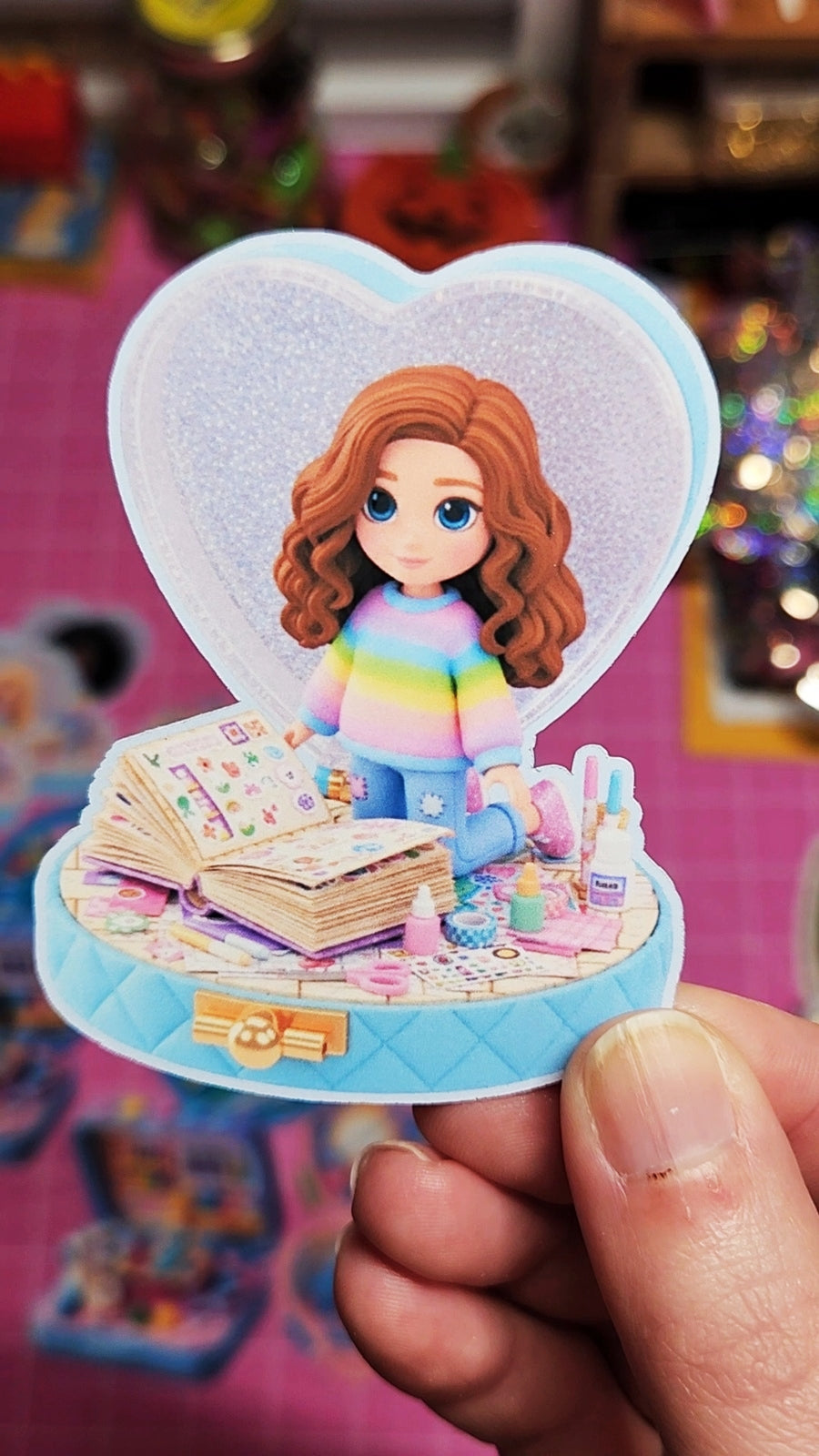 Toy figure of a girl with red hair in a heart-shaped box held by a hand against a colorful background.