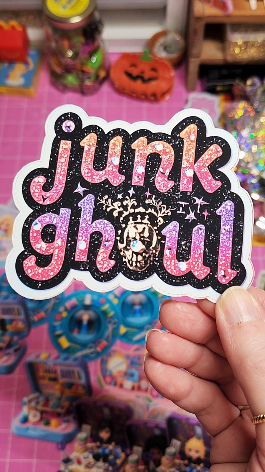 Sticker with 'junk ghoul' text held in front of a colorful, cluttered background