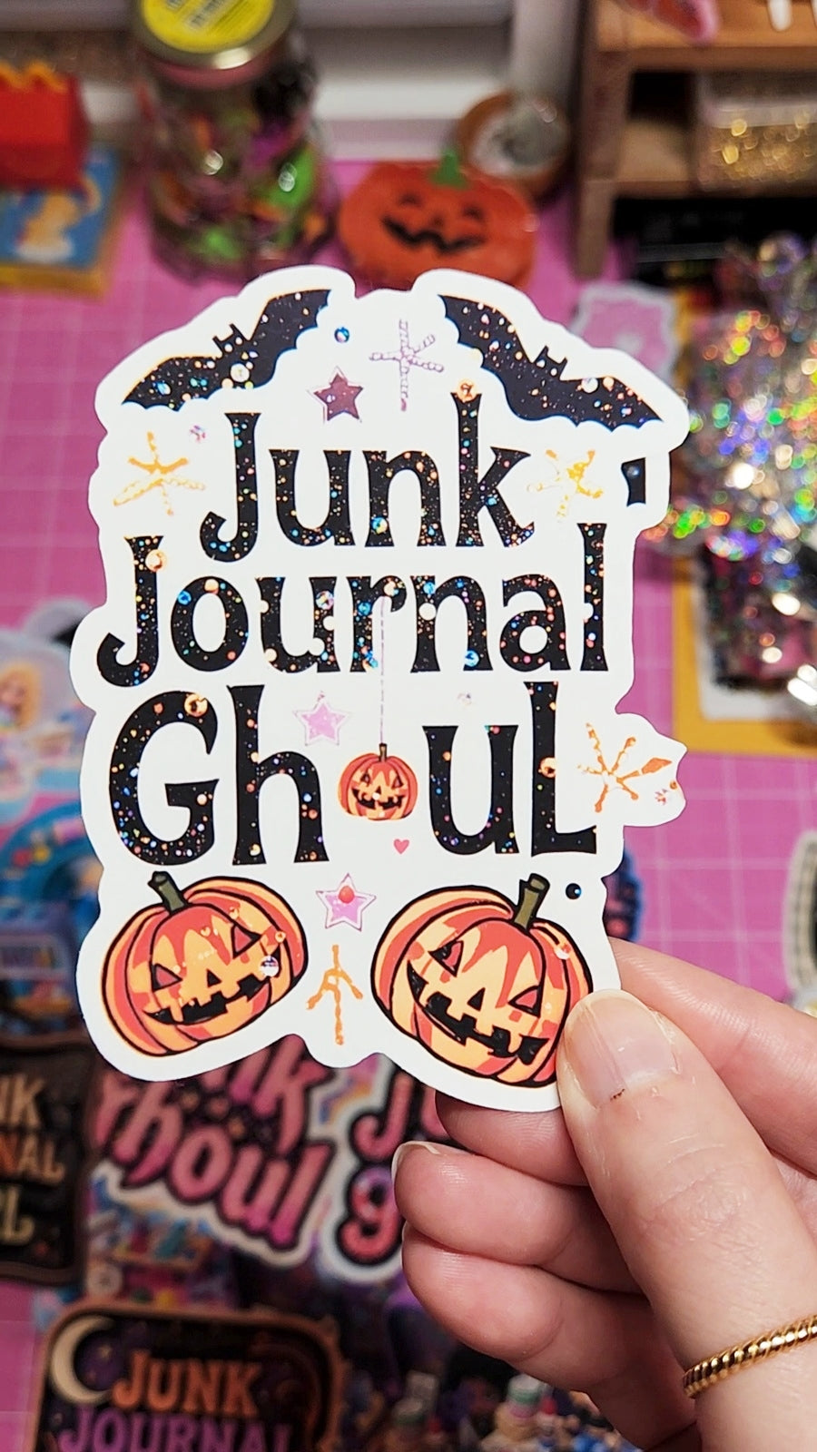 Sticker with 'Junk Journal Ghoul' text and pumpkin designs held by a hand against a colorful background.