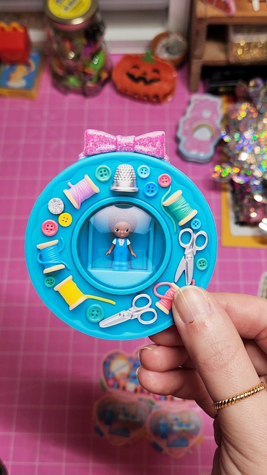 Toy sewing kit with a small doll figure held by a hand on a pink surface.