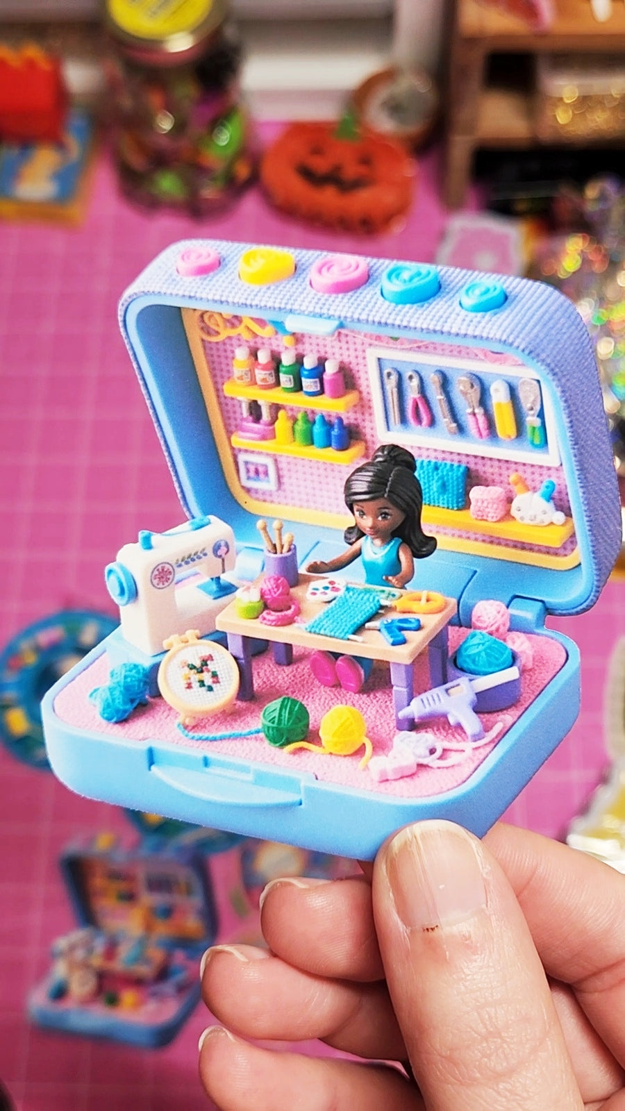 Toy sewing set with a doll inside a small box, held by a hand on a colorful background.