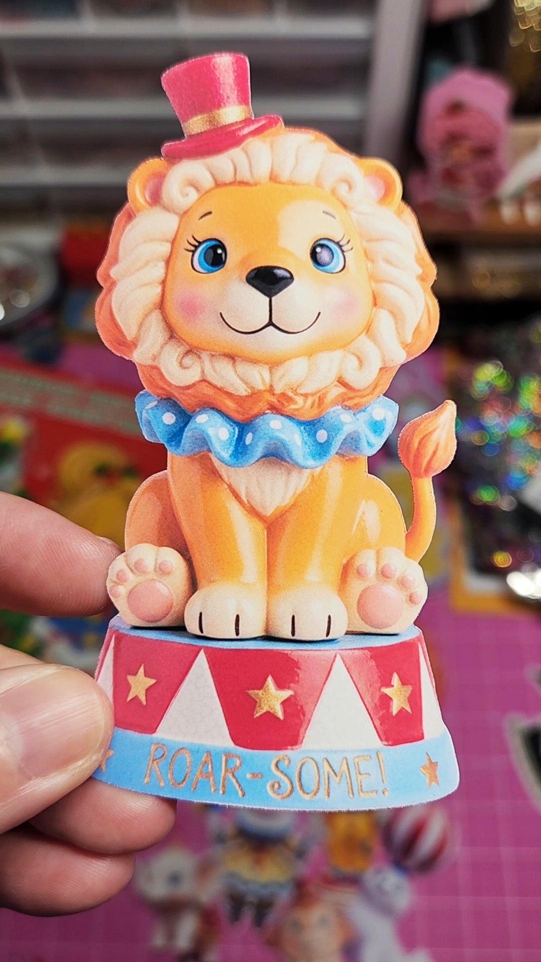 Colorful lion figurine with a clown hat and 'ROAR-SOME!' base held by a hand.