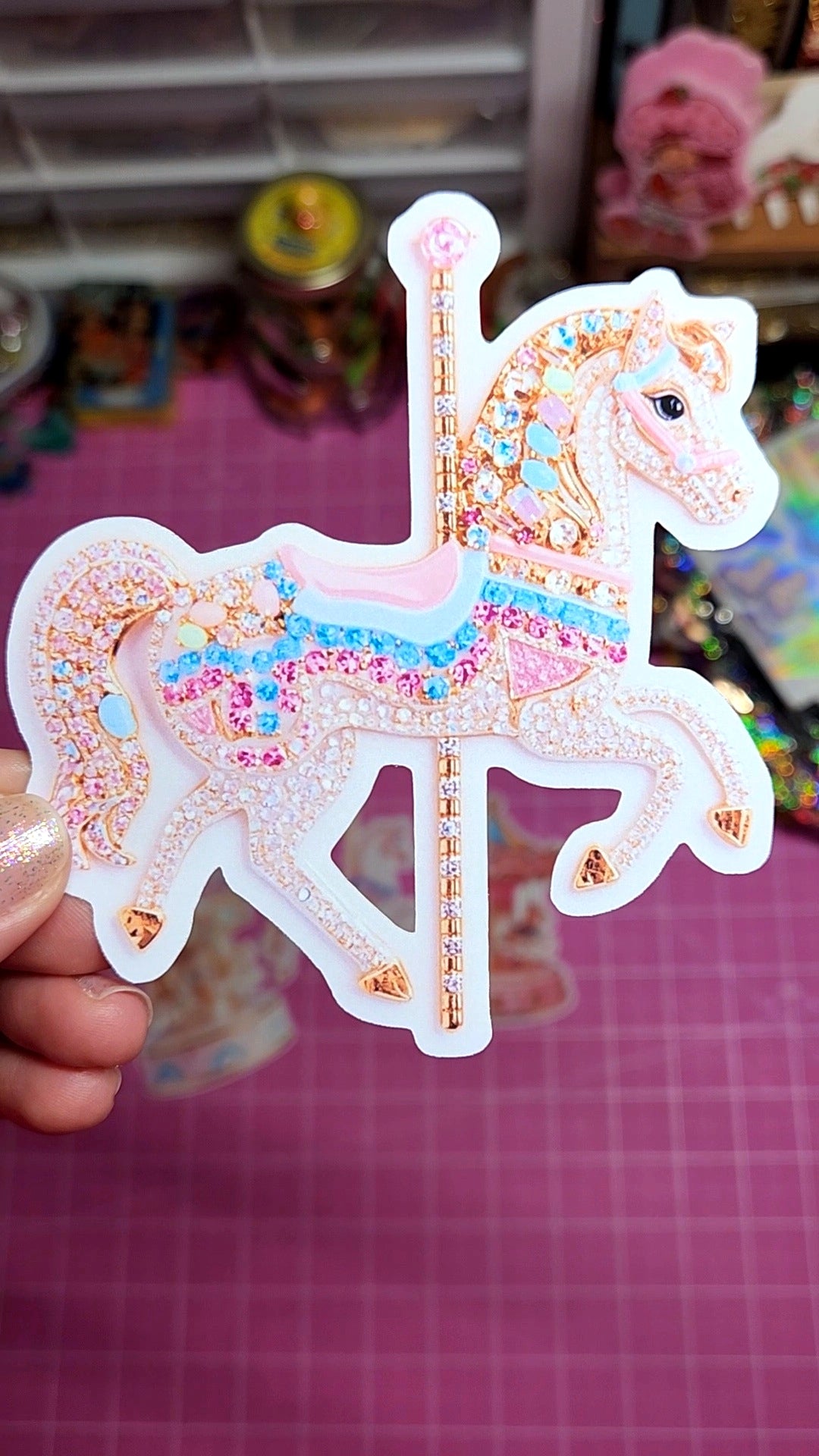 Decorative sticker of a carousel horse held by a hand on a pink checkered surface.