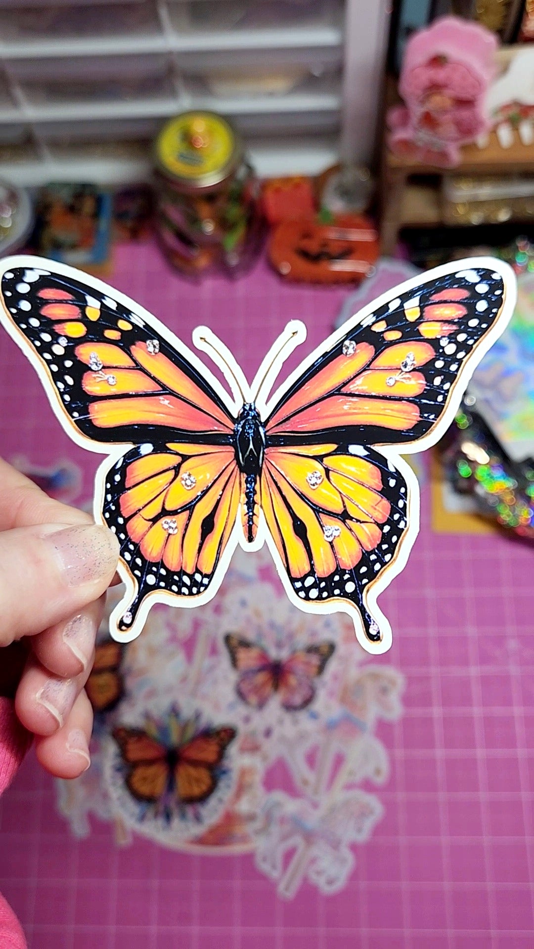 Butterfly-shaped sticker held by a hand on a pink surface with craft supplies in the background