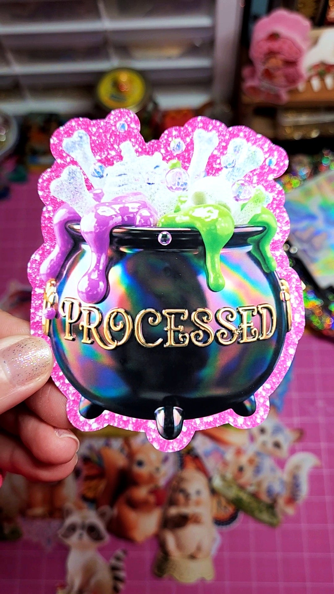 Colorful sticker with a cauldron and 'Processed' text, held over a blurred background with toys.
