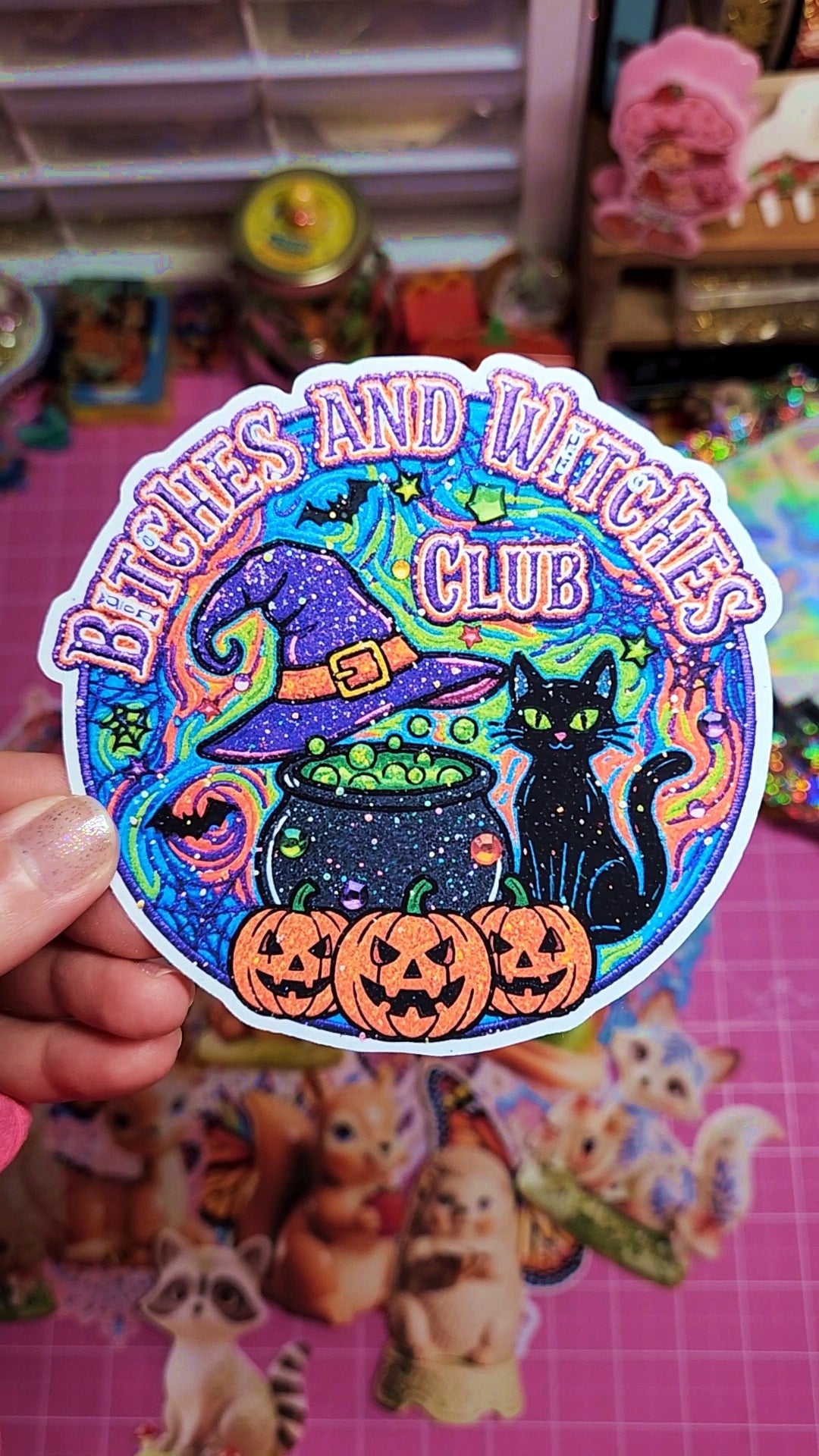 Sticker with 'Bitches and Witches Club' text, black cat, and pumpkins held in front of a colorful background.