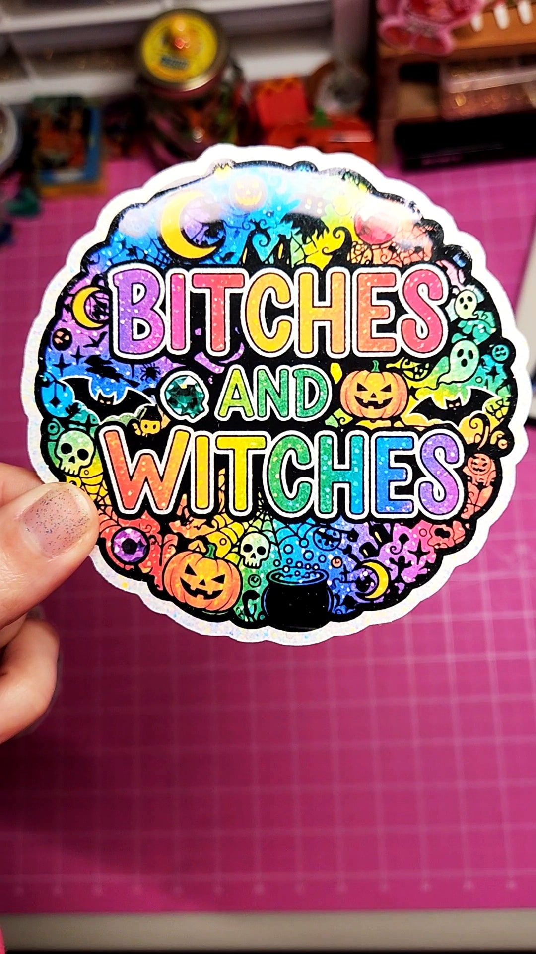 Sticker with colorful text 'Bitches and Witches' held over a pink grid background