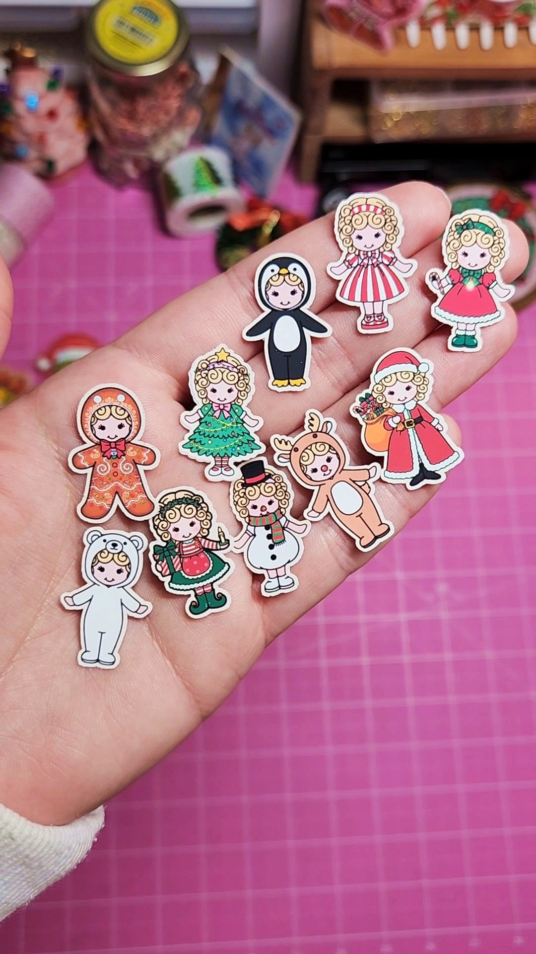 Hand holding a set of colorful cartoon character stickers on a pink grid background.