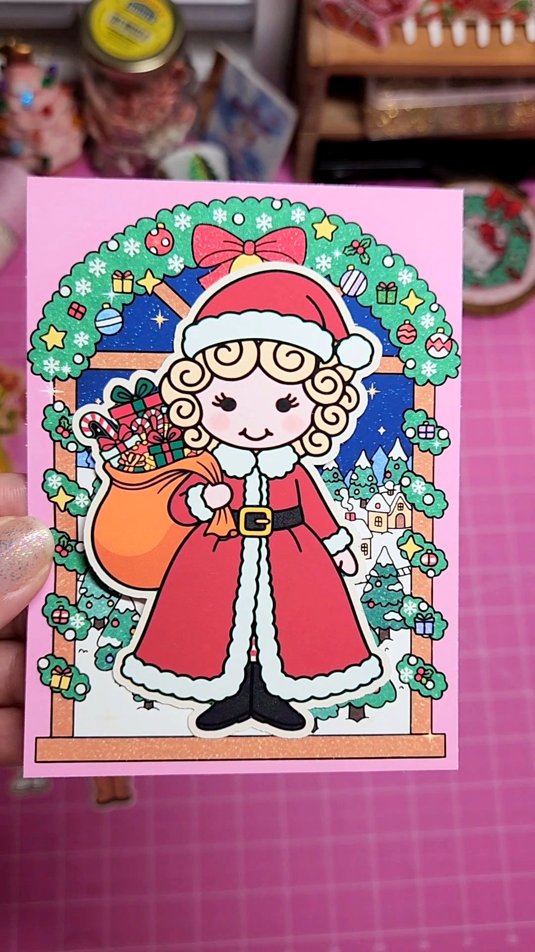 Illustration of a character in a Santa Claus outfit with festive decorations on a pink card.