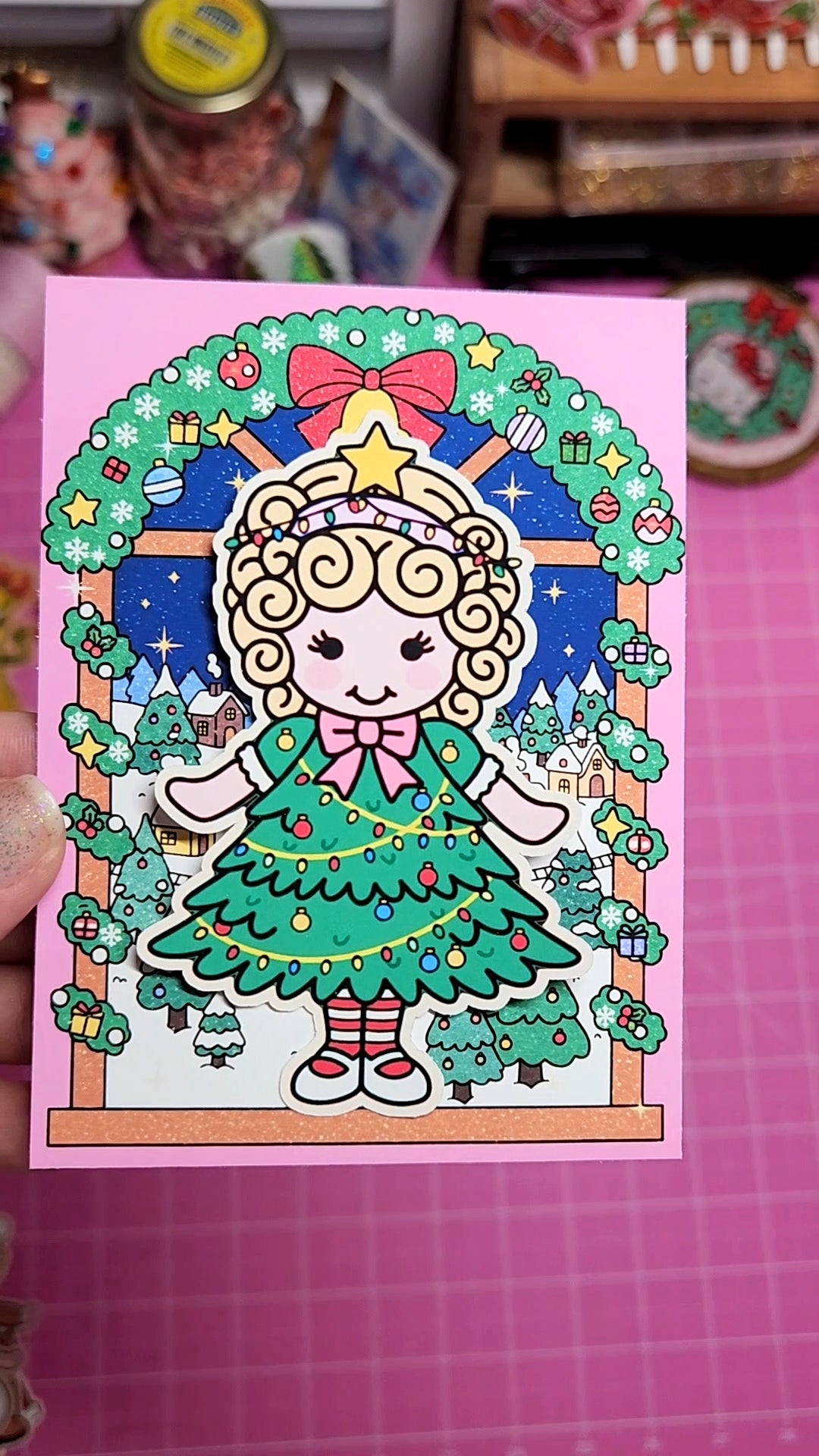 Illustration of a character in a green dress with a Christmas tree on a pink card.