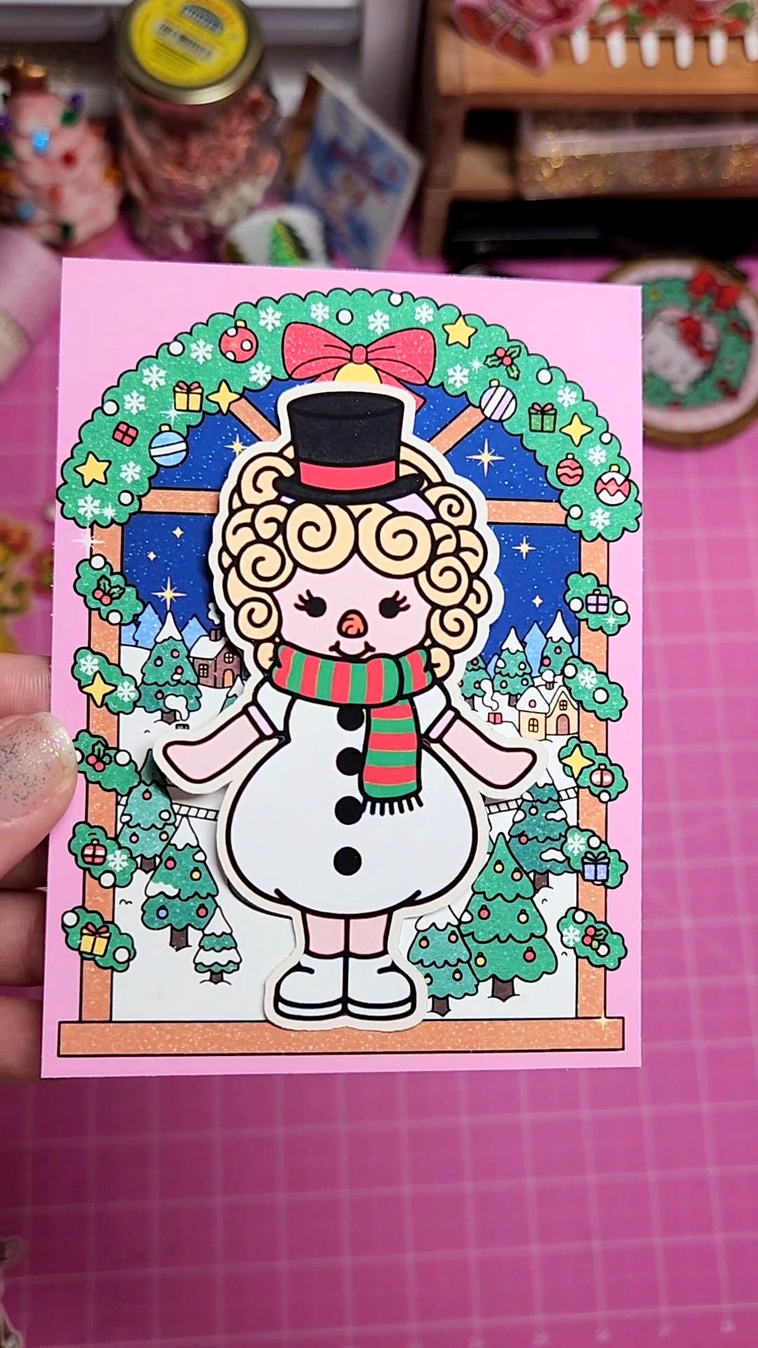 Illustrated snowman character on a pink card with festive decorations.