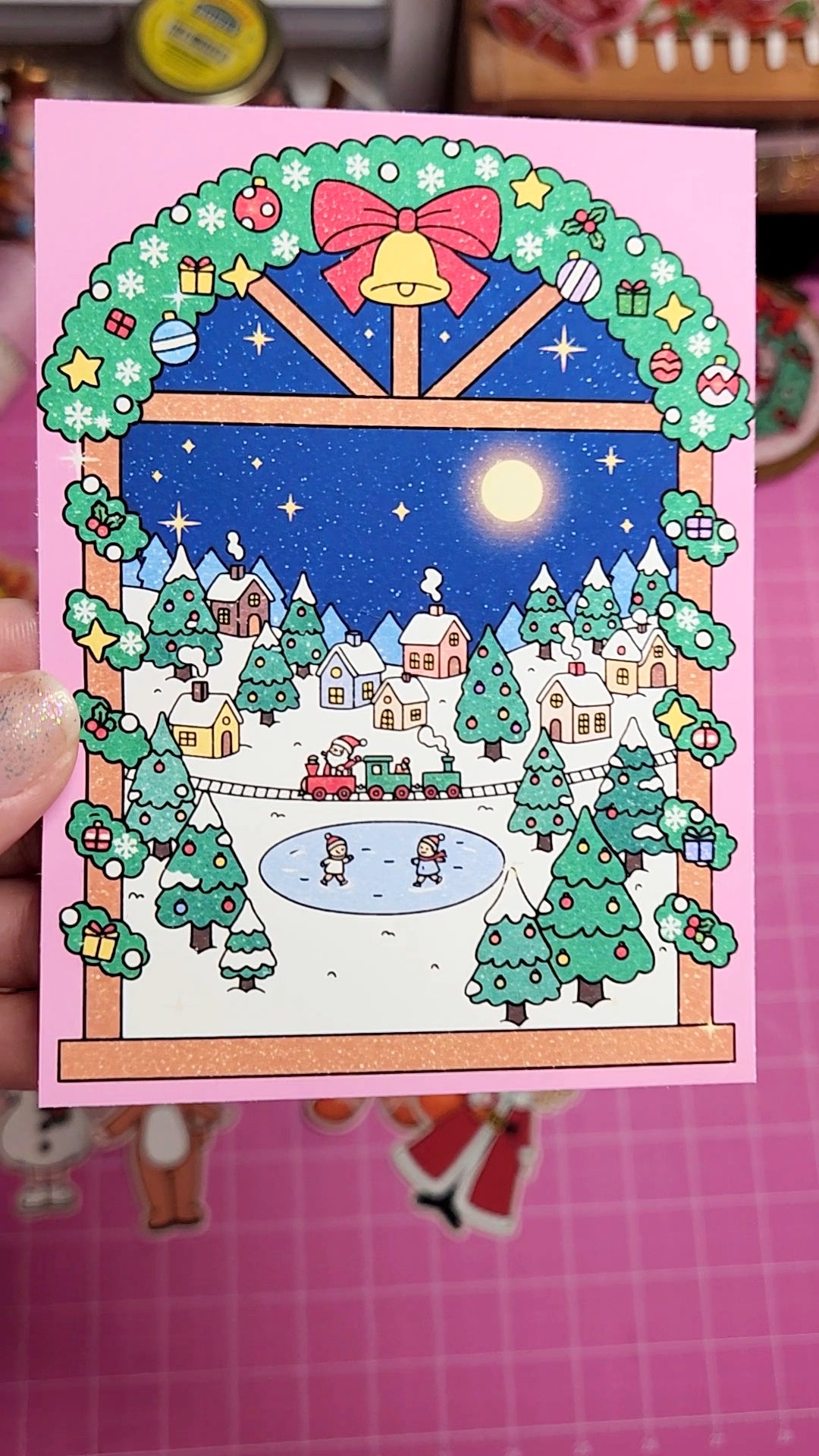 Christmas-themed card with a snowy village scene through a window, held by a hand on a pink background.