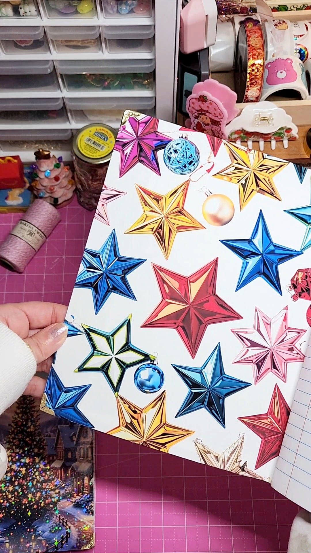 Colorful star-shaped decorations on a white card held by a hand, with a craft room in the background.