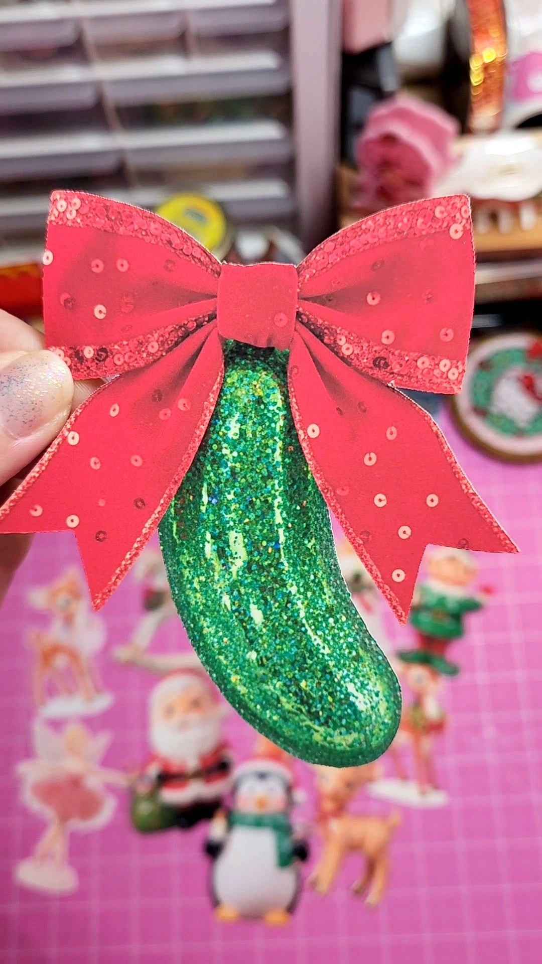 Green glittery pickle with a red bow on a pink background with Christmas-themed decorations.