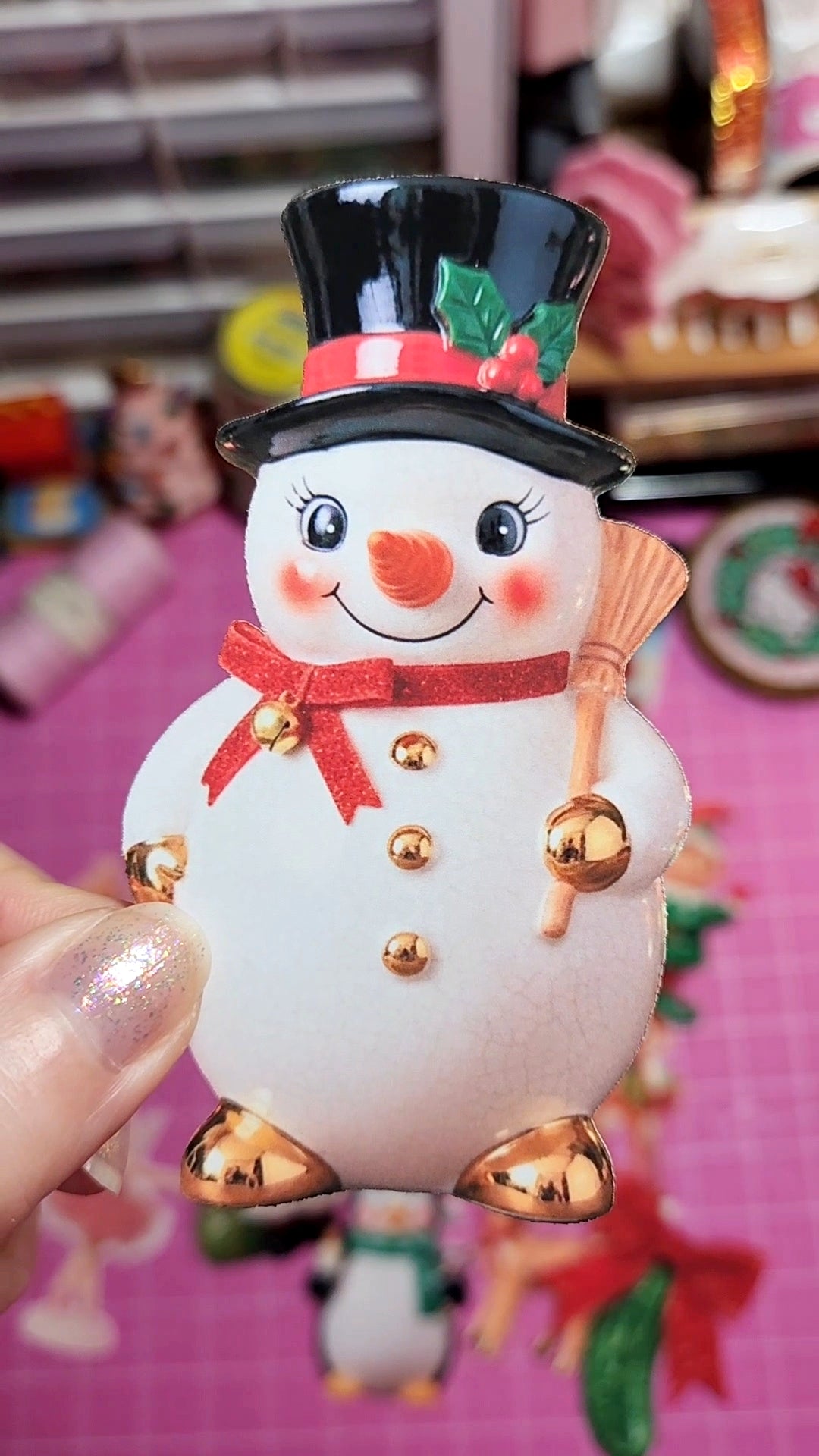 Decorative snowman figurine with a black top hat and gold jingle bells on a pink background.
