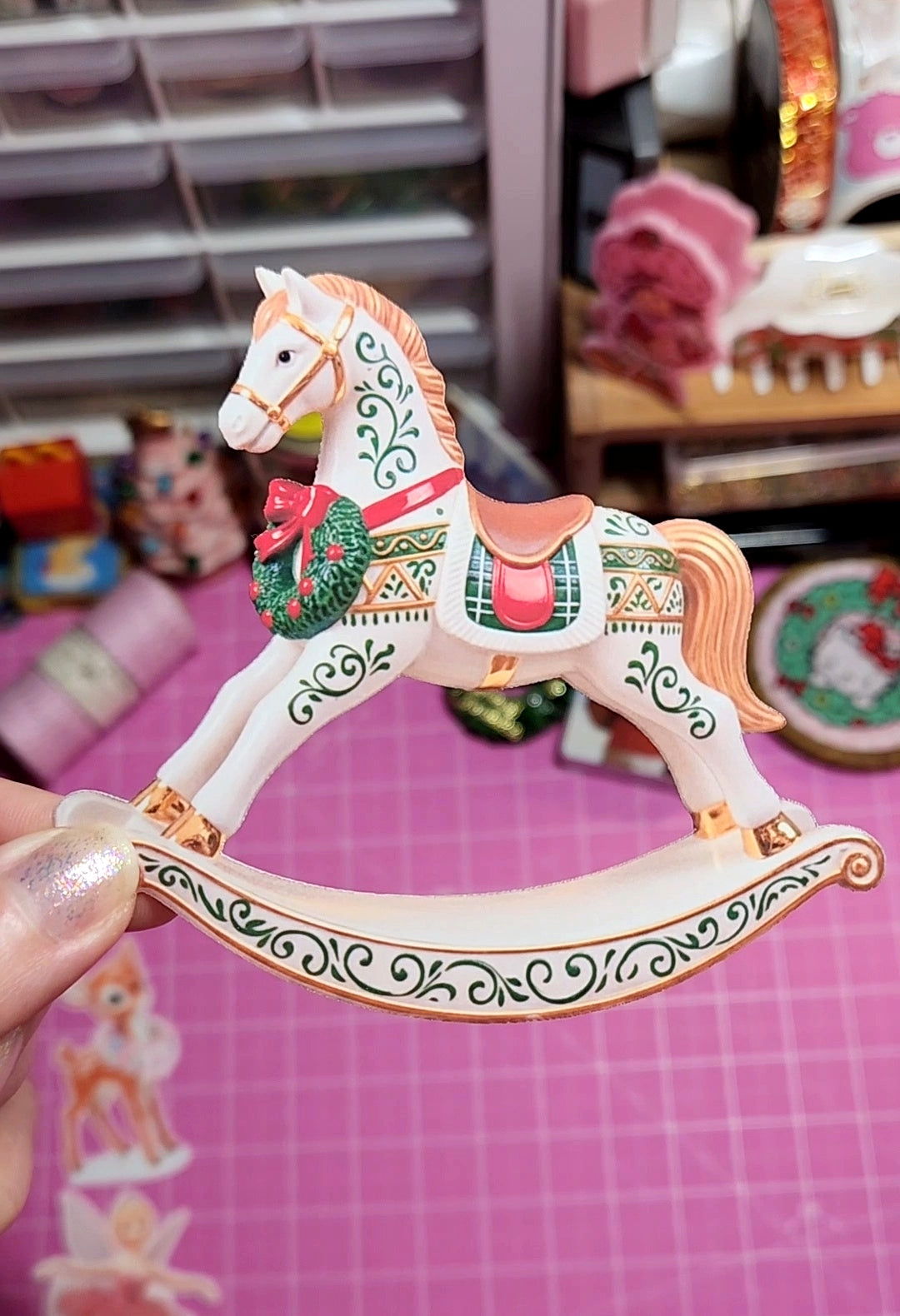 Decorative rocking horse toy held by a hand on a pink surface with craft materials in the background.