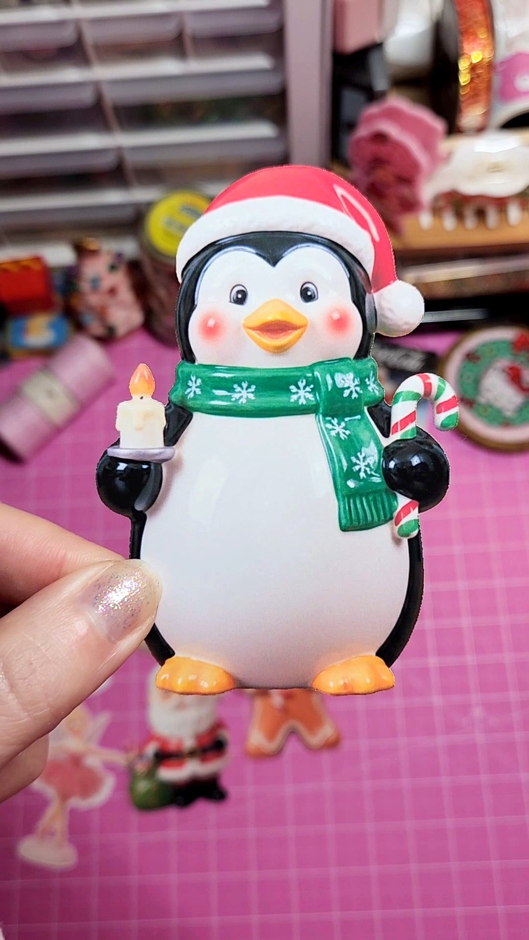 Penguin figurine with a Santa hat, green scarf, and candy cane on a pink background.