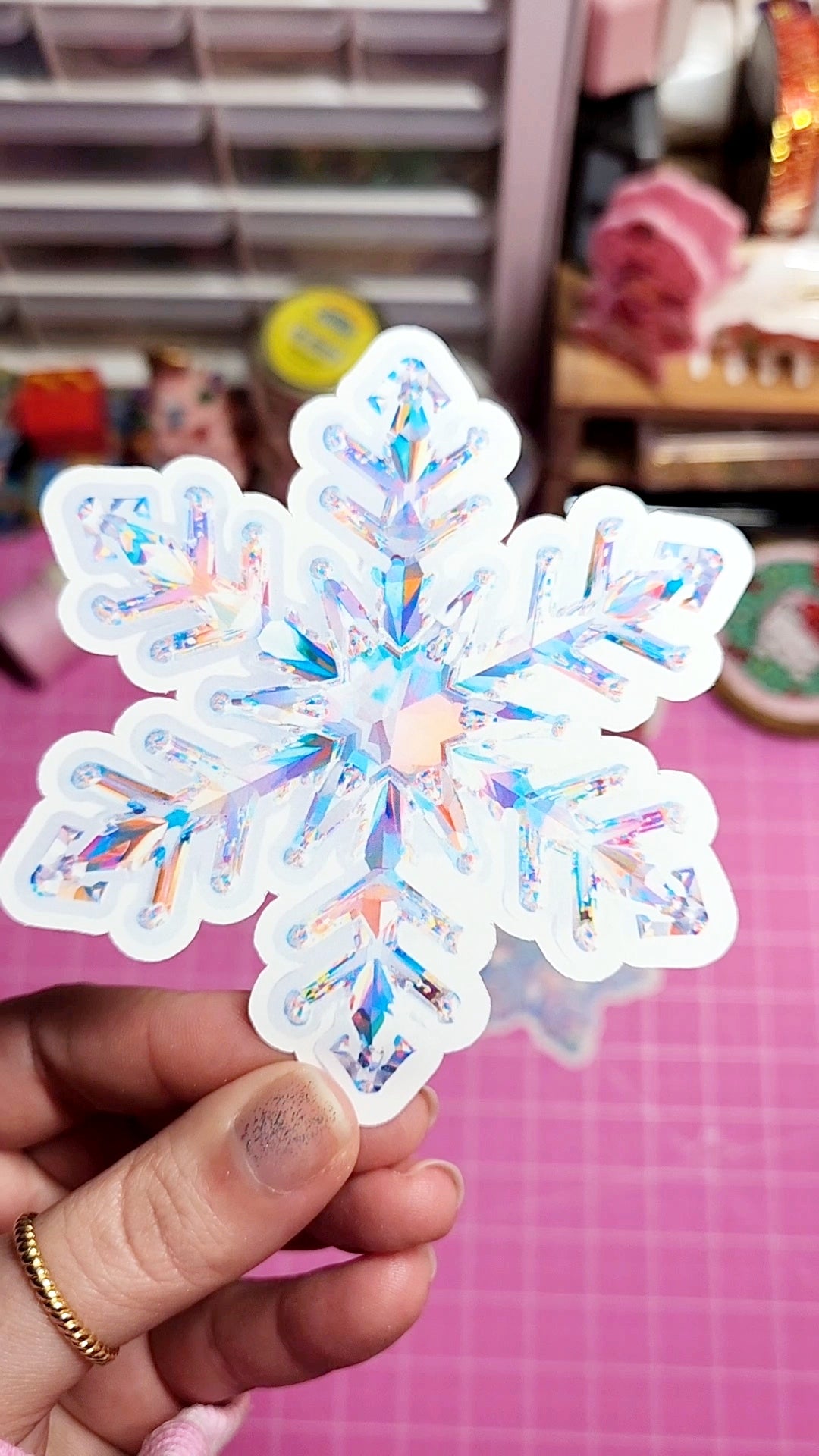 Iridescent snowflake-shaped sticker held by a hand on a pink surface with blurred background