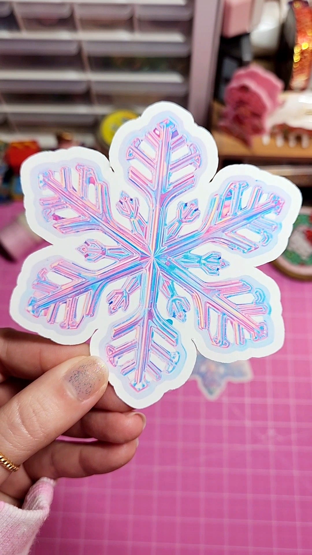 Holographic snowflake sticker held by a hand on a pink cutting mat.