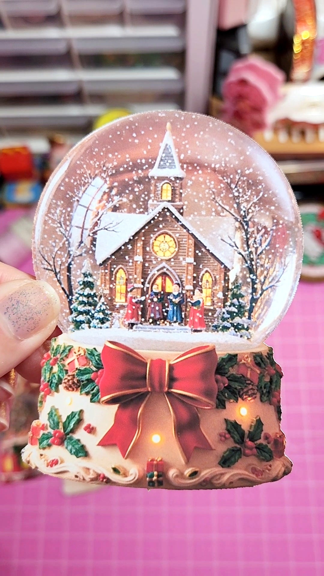 Christmas-themed snow globe with a church scene inside, held by a hand on a pink surface.