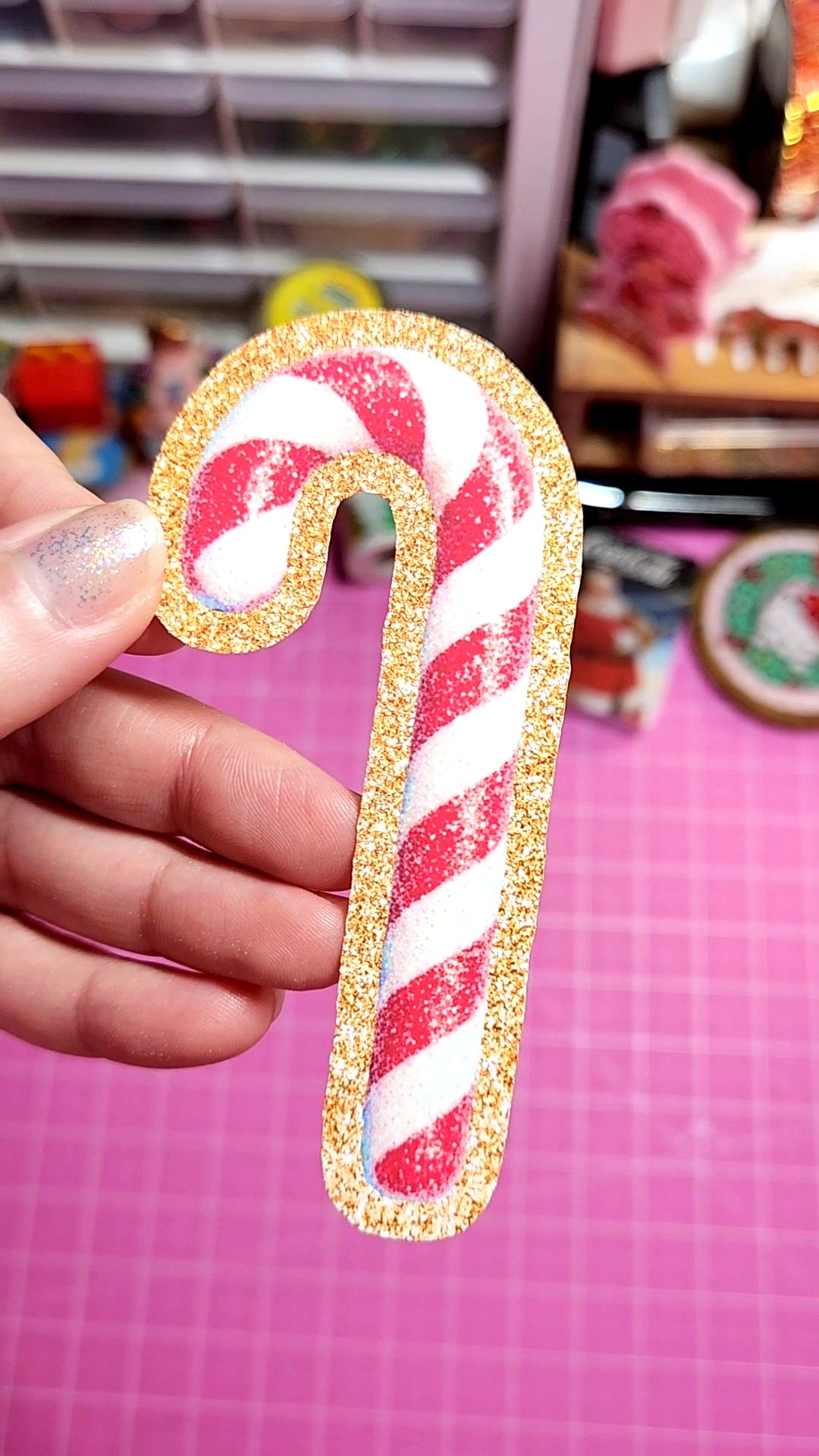 Candy cane-shaped decorative item with glittery edge held by a hand on a pink background.