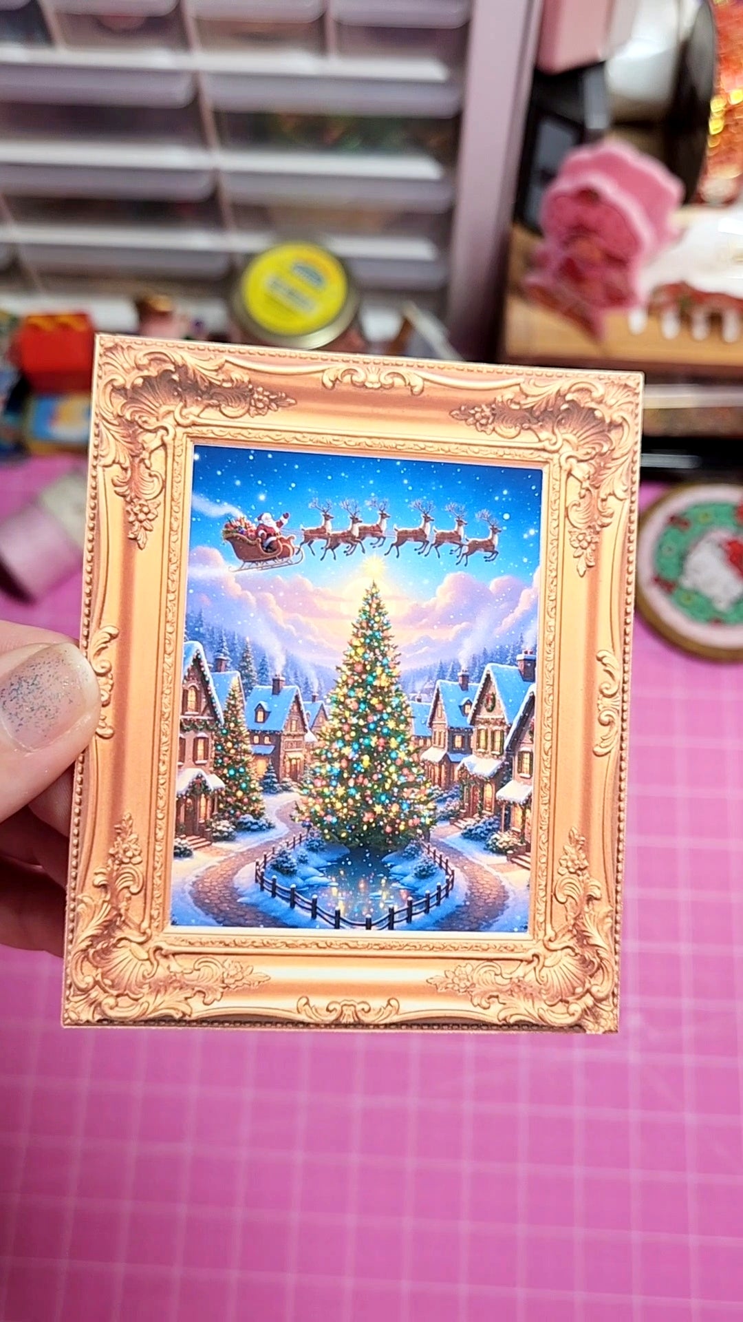 Decorative Christmas scene with Santa's sleigh and reindeer in a gold frame on a pink surface.