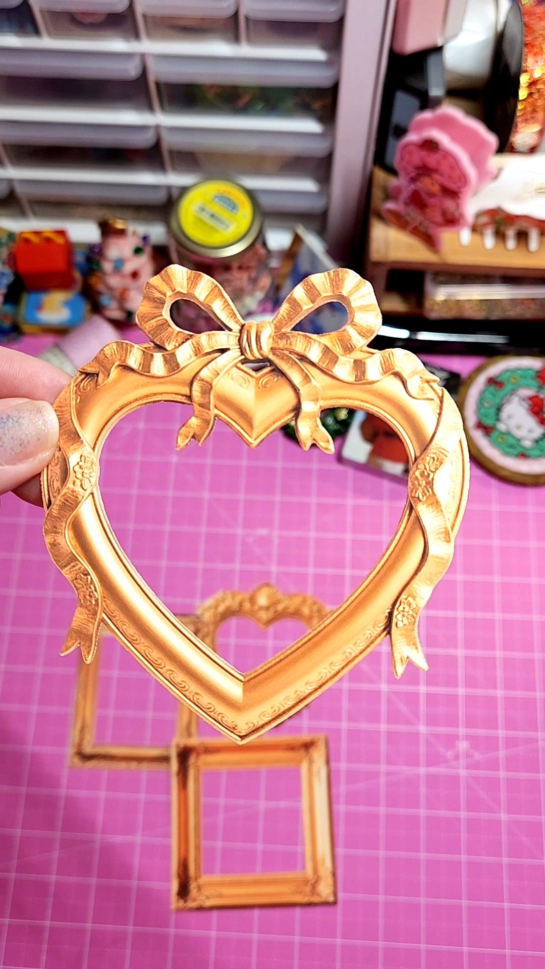 Gold heart-shaped frame with bow design held by a hand on a pink grid background.