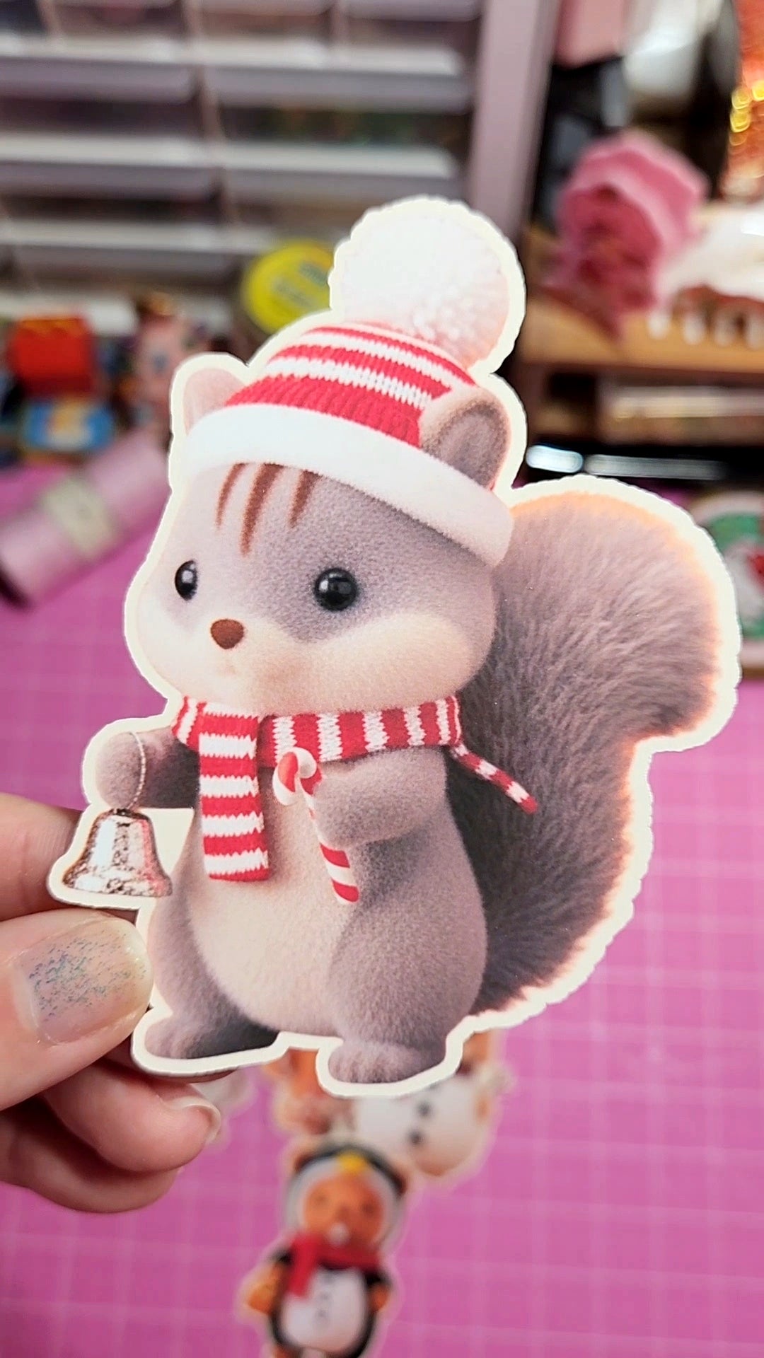 Plush toy squirrel with a red and white striped hat and scarf held by a hand on a pink background.