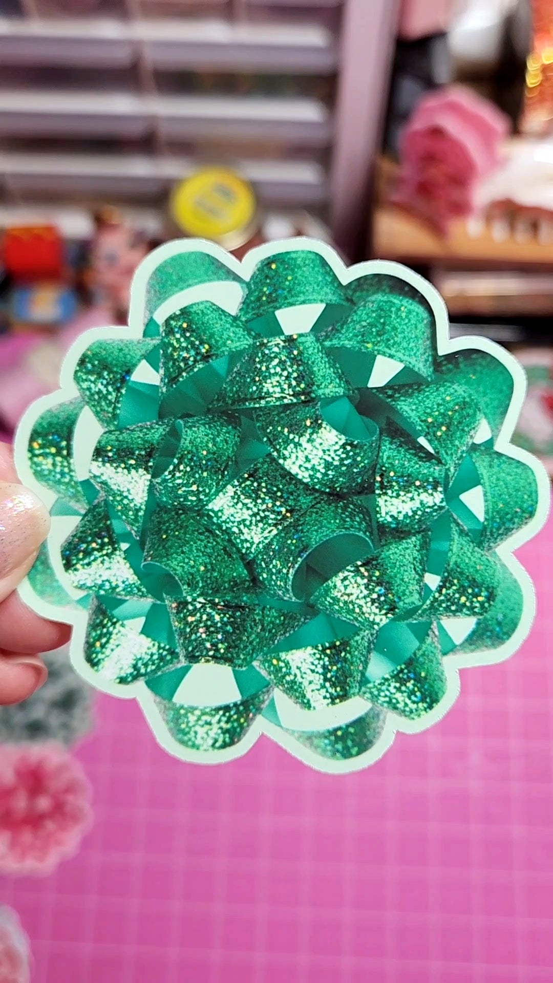 Green glittery bow-shaped object held by a hand against a pink background
