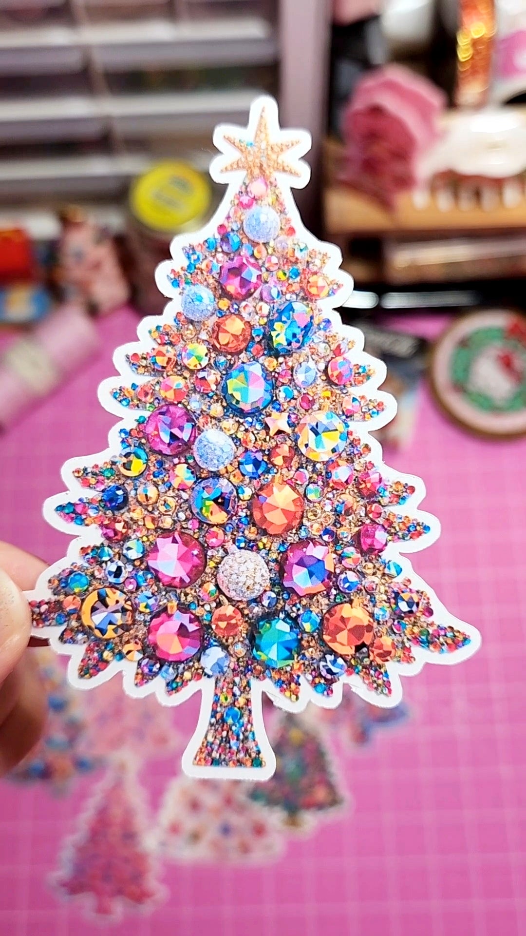 Colorful Christmas tree-shaped decorative item with rhinestones on a pink surface.