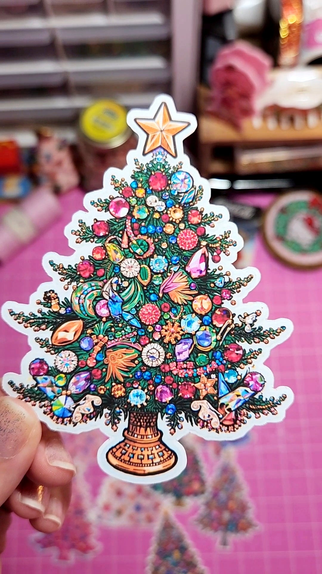 Colorful Christmas tree sticker with decorative elements held by a hand against a blurred indoor background.