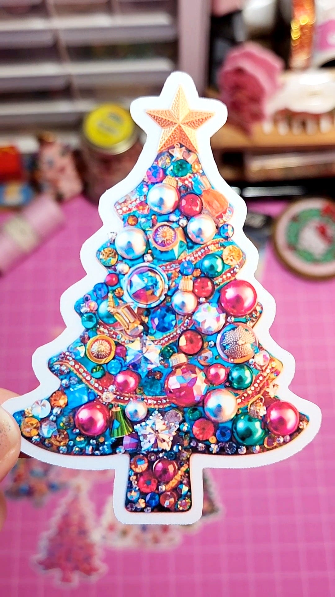 Colorful Christmas tree-shaped decoration with beads and sequins held by a hand against a blurred indoor background.
