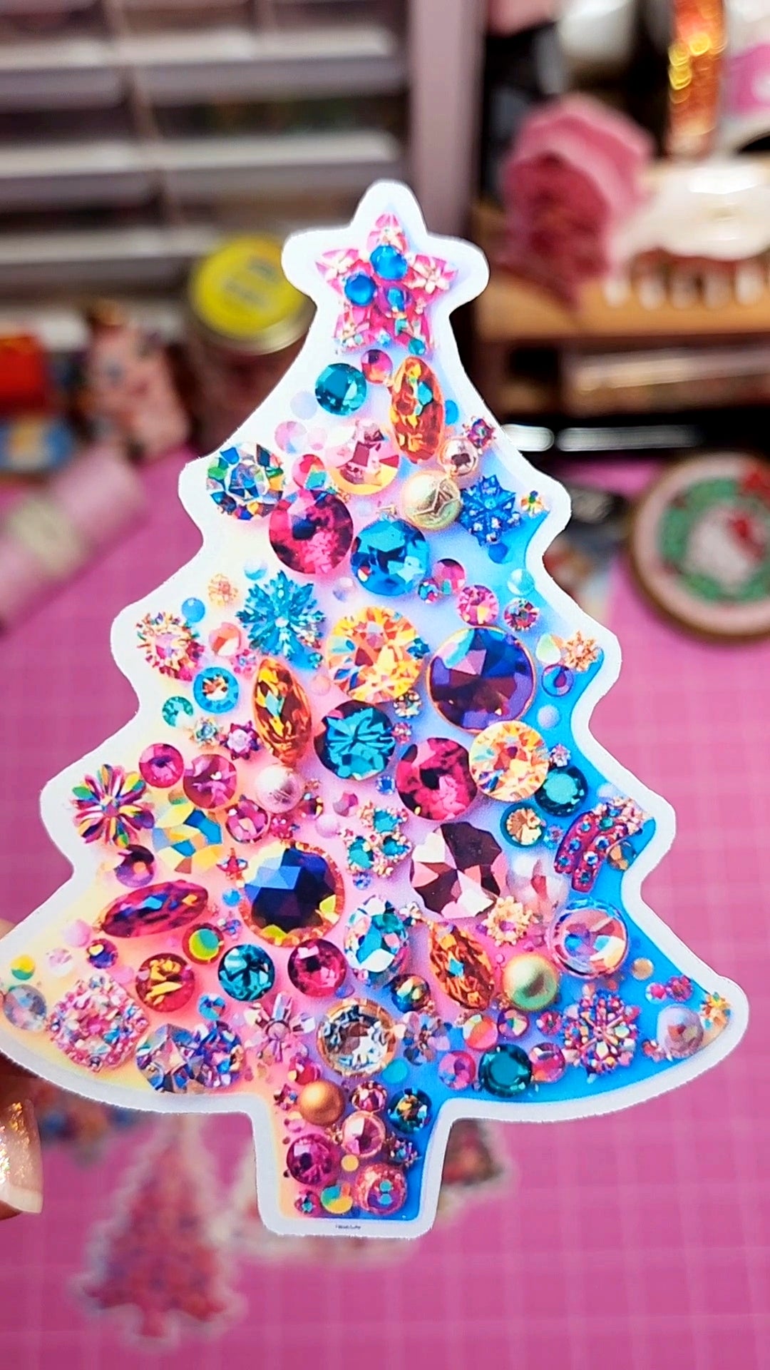 Colorful Christmas tree-shaped sticker with rhinestones on a pink surface.