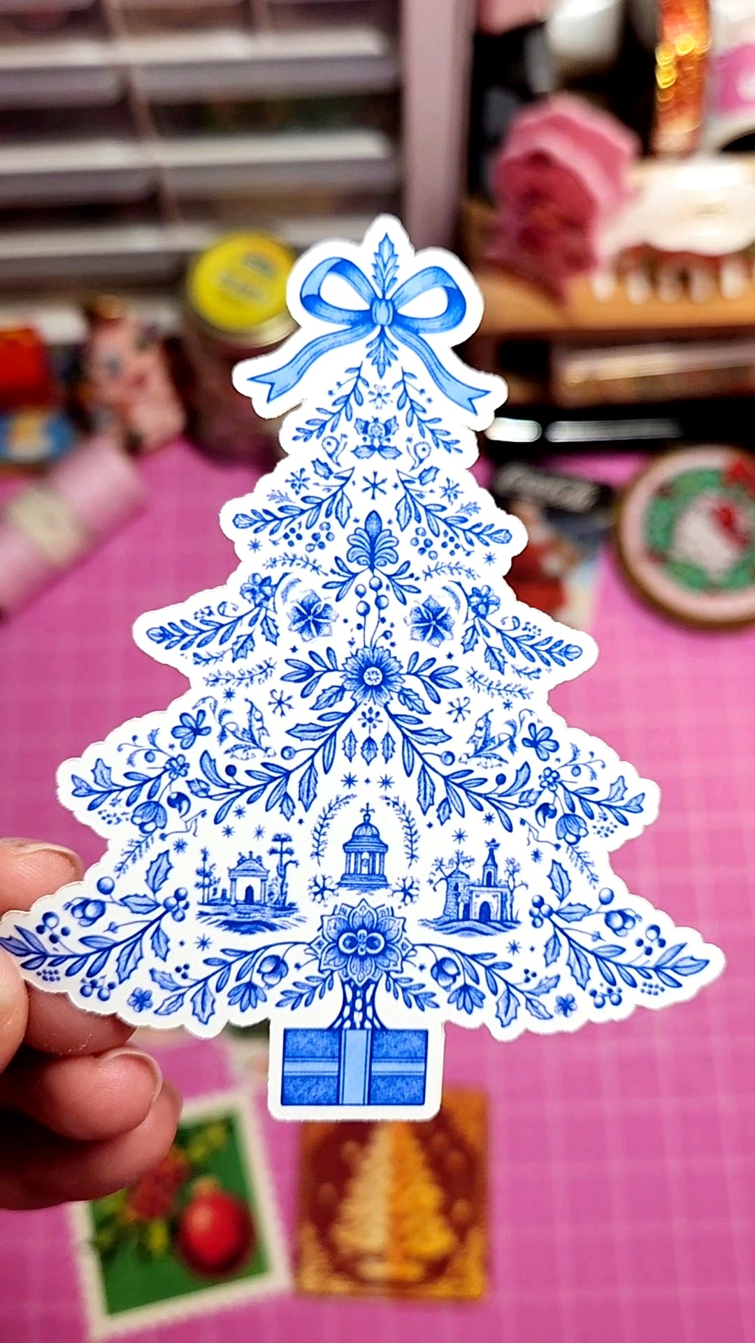 Decorative Christmas tree sticker with blue patterns held by a hand on a pink surface.