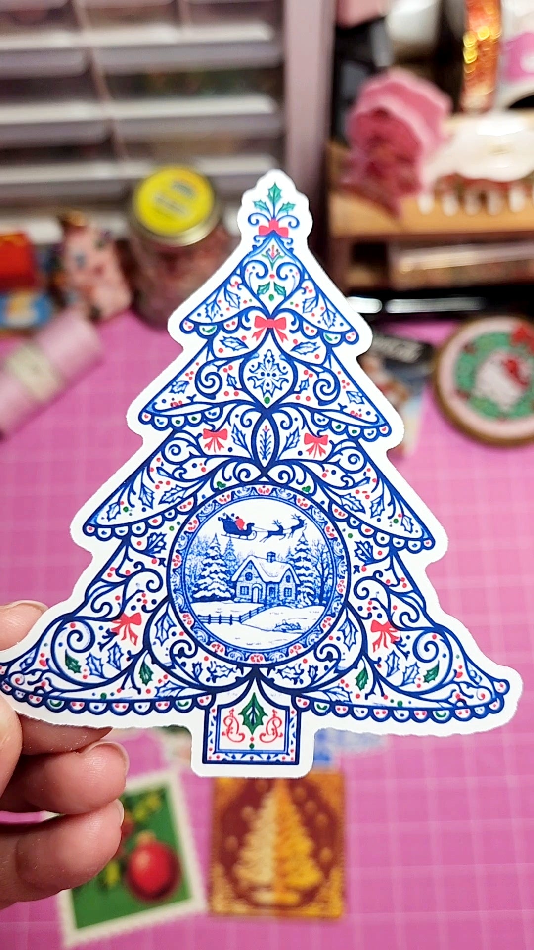 Decorative Christmas tree sticker held by a hand on a pink surface with blurred background