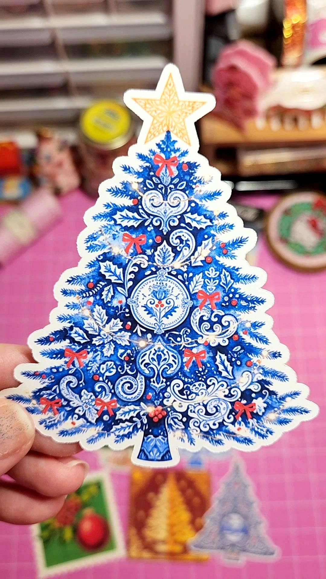 Decorative Christmas tree sticker with blue and white design held by a hand against a blurred indoor background.