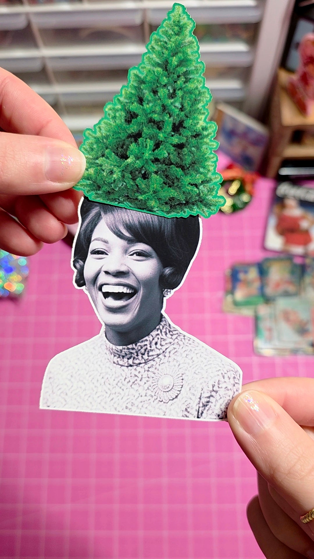 Person holding a paper cutout of a smiling woman with a green tree on her head against a pink grid background.
