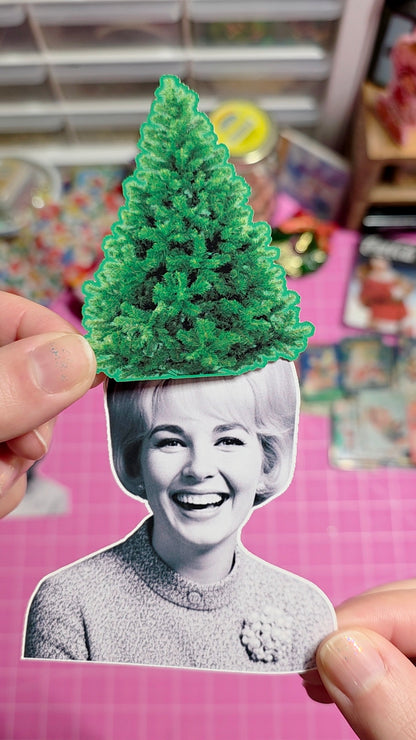 Person holding a cutout of a smiling woman with a green Christmas tree on her head against a pink background.