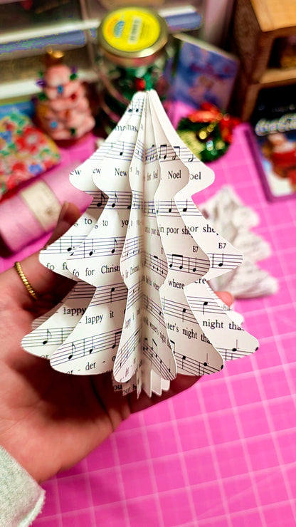 Hand holding a paper Christmas tree with sheet music design on a pink surface.