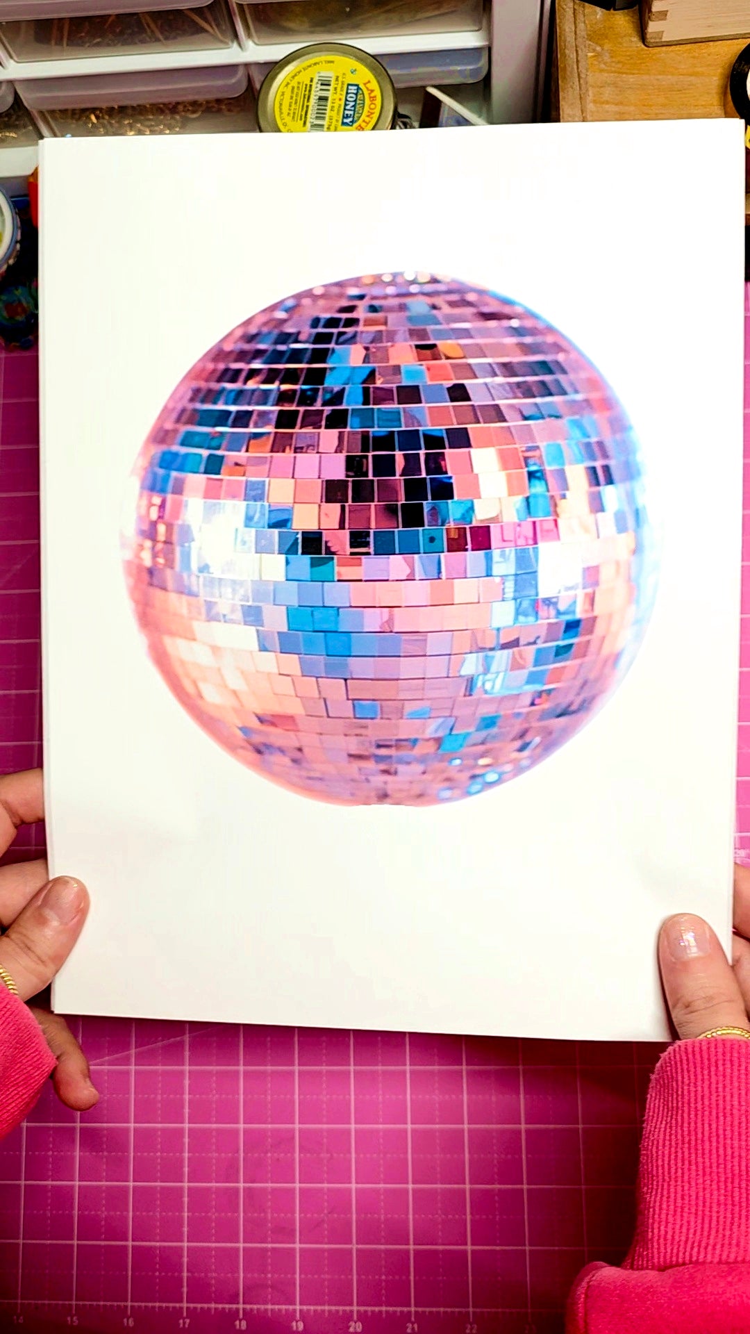Colorful disco ball on a white sheet held by hands on a pink cutting mat.