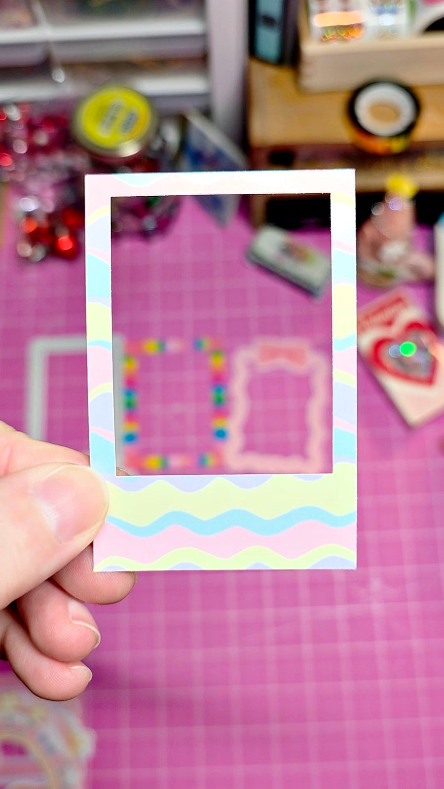 Hand holding a colorful photo frame with a pink background against a blurred indoor setting.