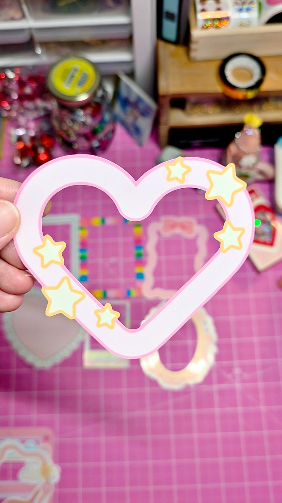 Heart-shaped frame with star decorations held by a hand on a pink checkered background.