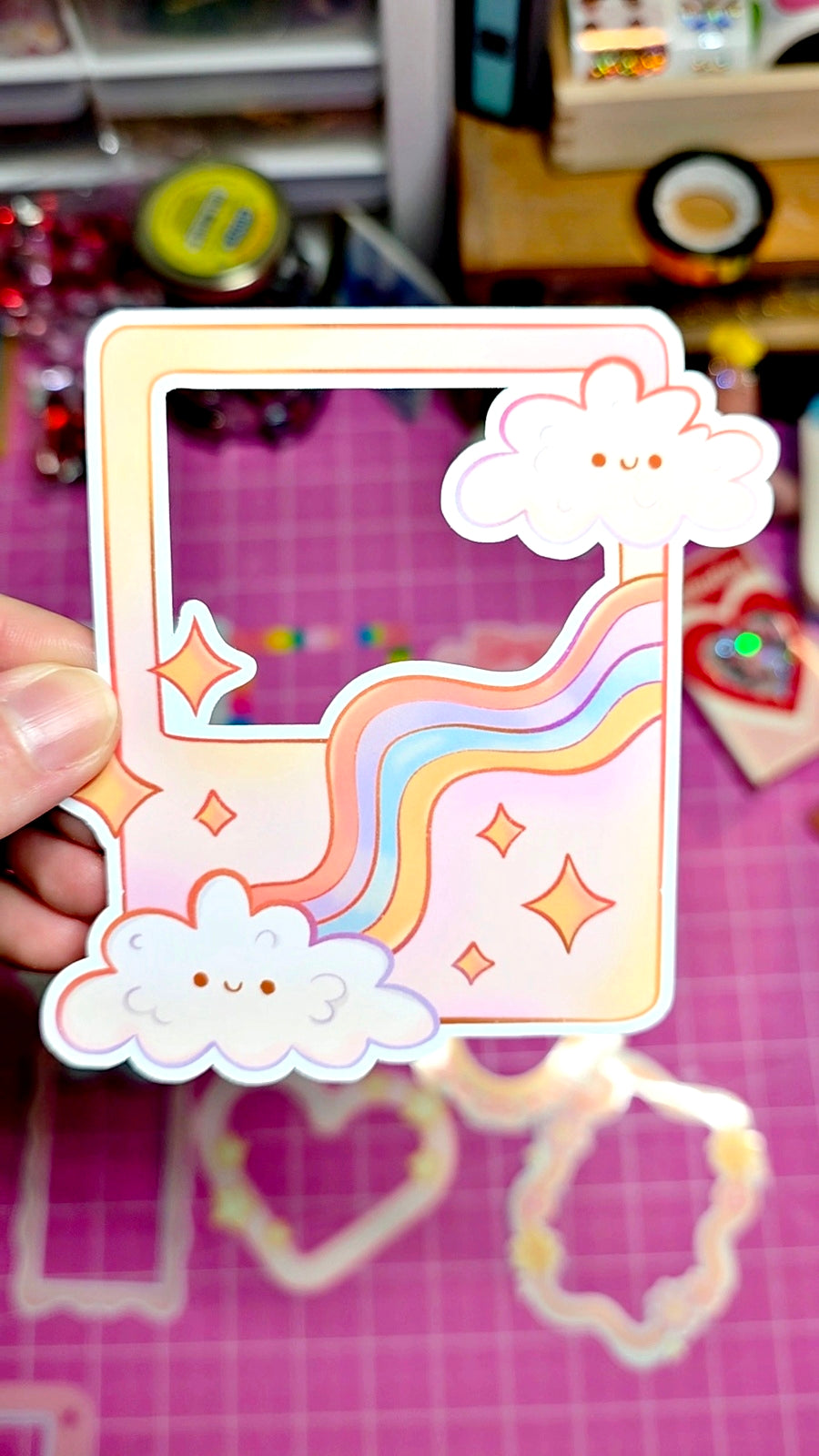 Decorative phone case with cloud and rainbow design held by a hand on a pink surface.