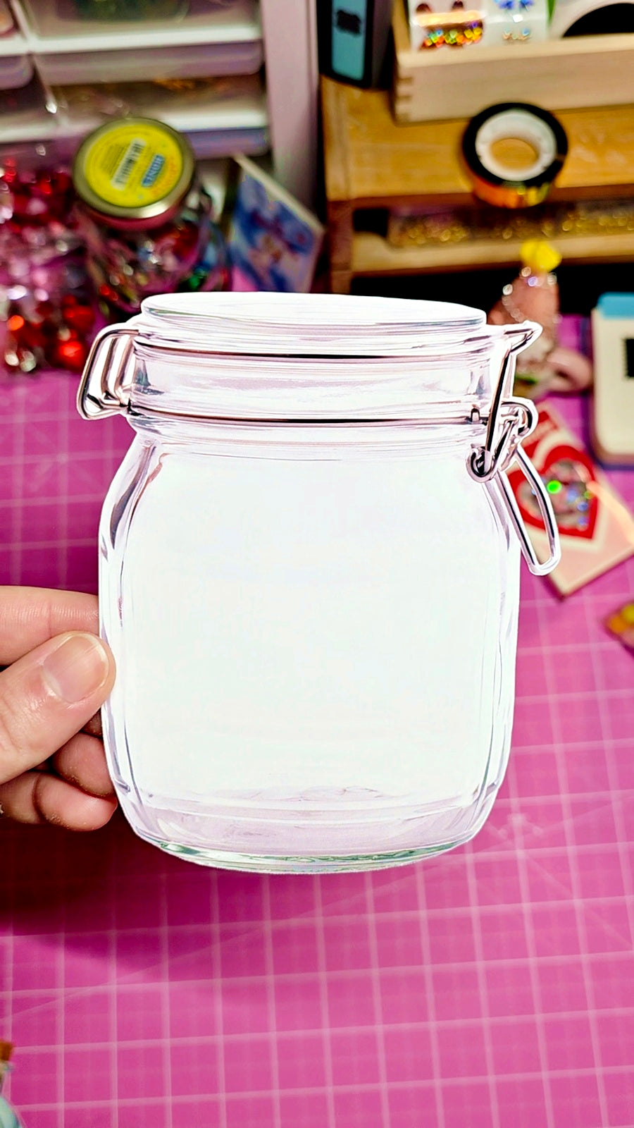 Clear glass jar with a lid held by a hand on a pink surface with toys in the background