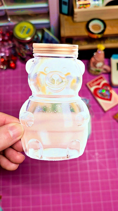 Clear plastic bottle with bear design held by a hand on a pink surface with toys in the background