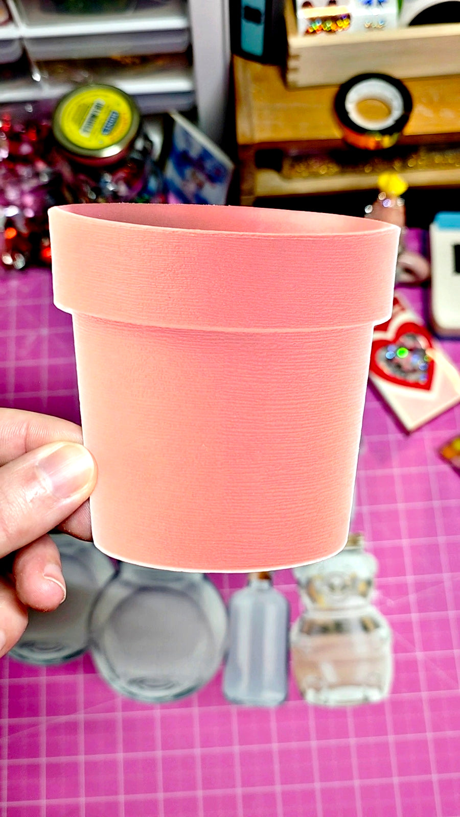 Pink cup held by a hand on a pink checkered surface with various items in the background