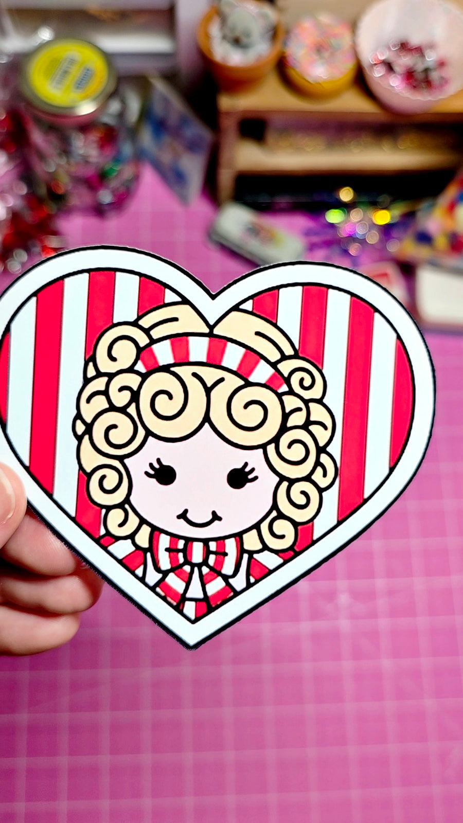Heart-shaped sticker with a cartoon character held by a hand on a pink checkered background.